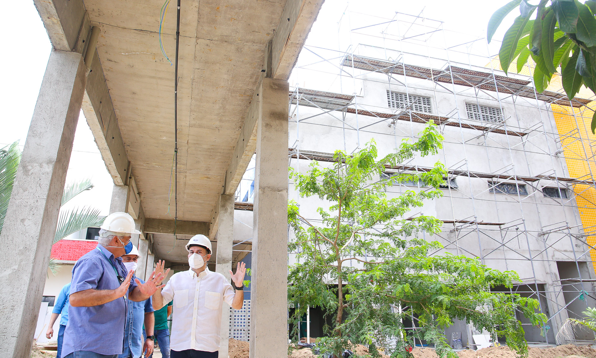 prefeito no canto inferior esquerdo de máscara usando capacete de engenheiro conversando com senhor também de máscara e capacete, e hospital em obras ao fundo